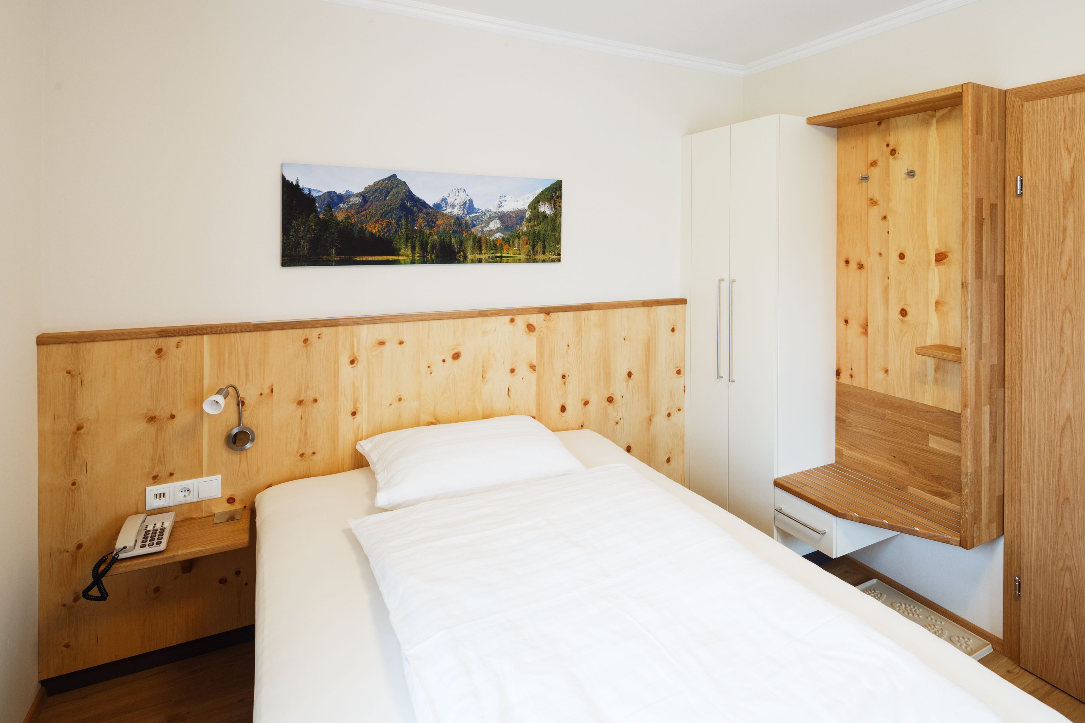 A bedroom with a wooden headboard, a bed with white bedding, a bedside telephone, a wooden cabinet with a built-in shelf, and a framed picture of a mountain landscape on the wall.