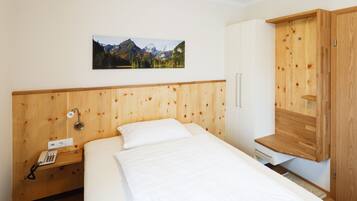 A bedroom with a wooden headboard, a bed with white bedding, a bedside telephone, a wooden cabinet with a built-in shelf, and a framed picture of a mountain landscape on the wall.