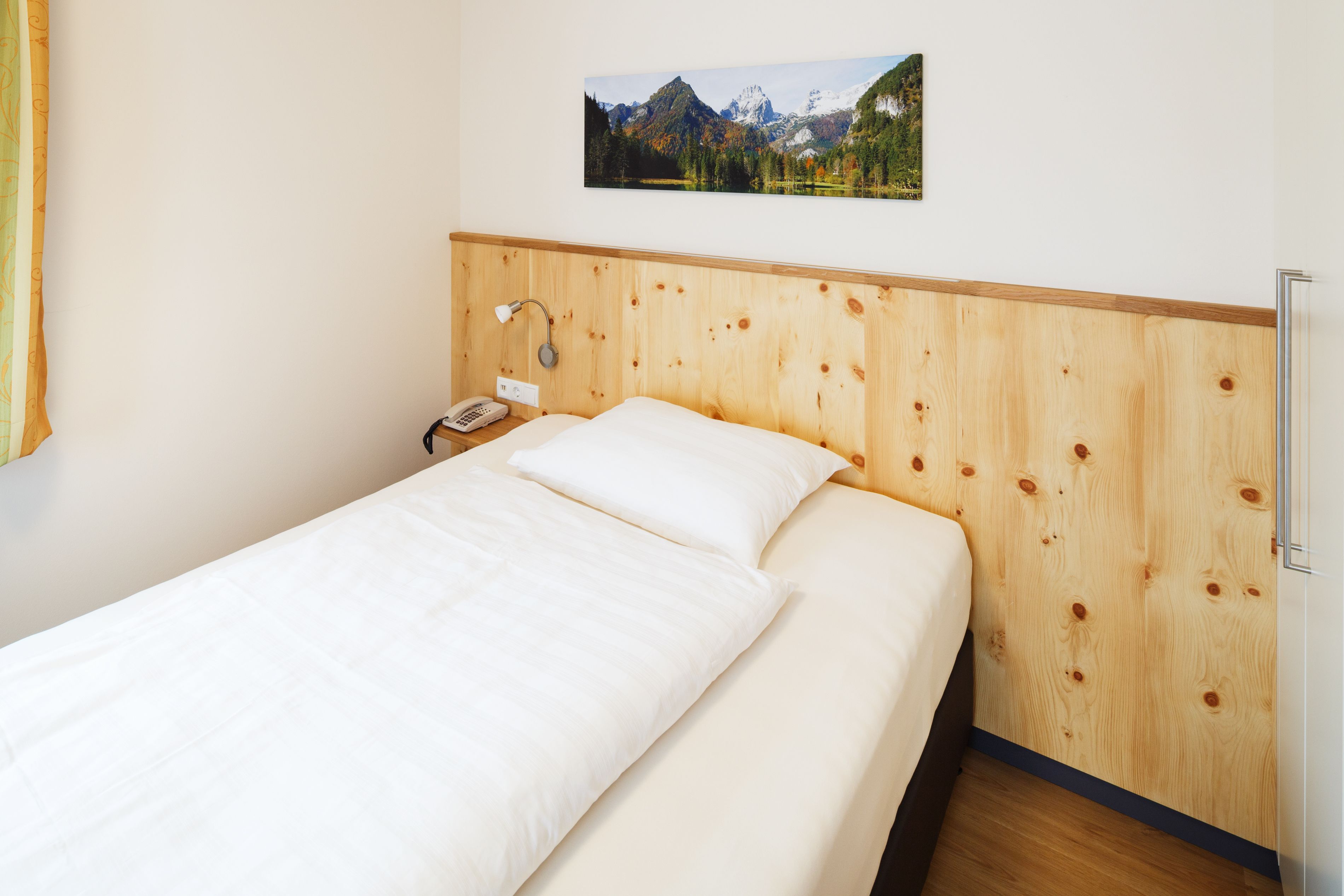 A hotel room with a wooden headboard, a bed with white linens, a nightstand with a lamp, and a framed picture of a mountain landscape on the wall.