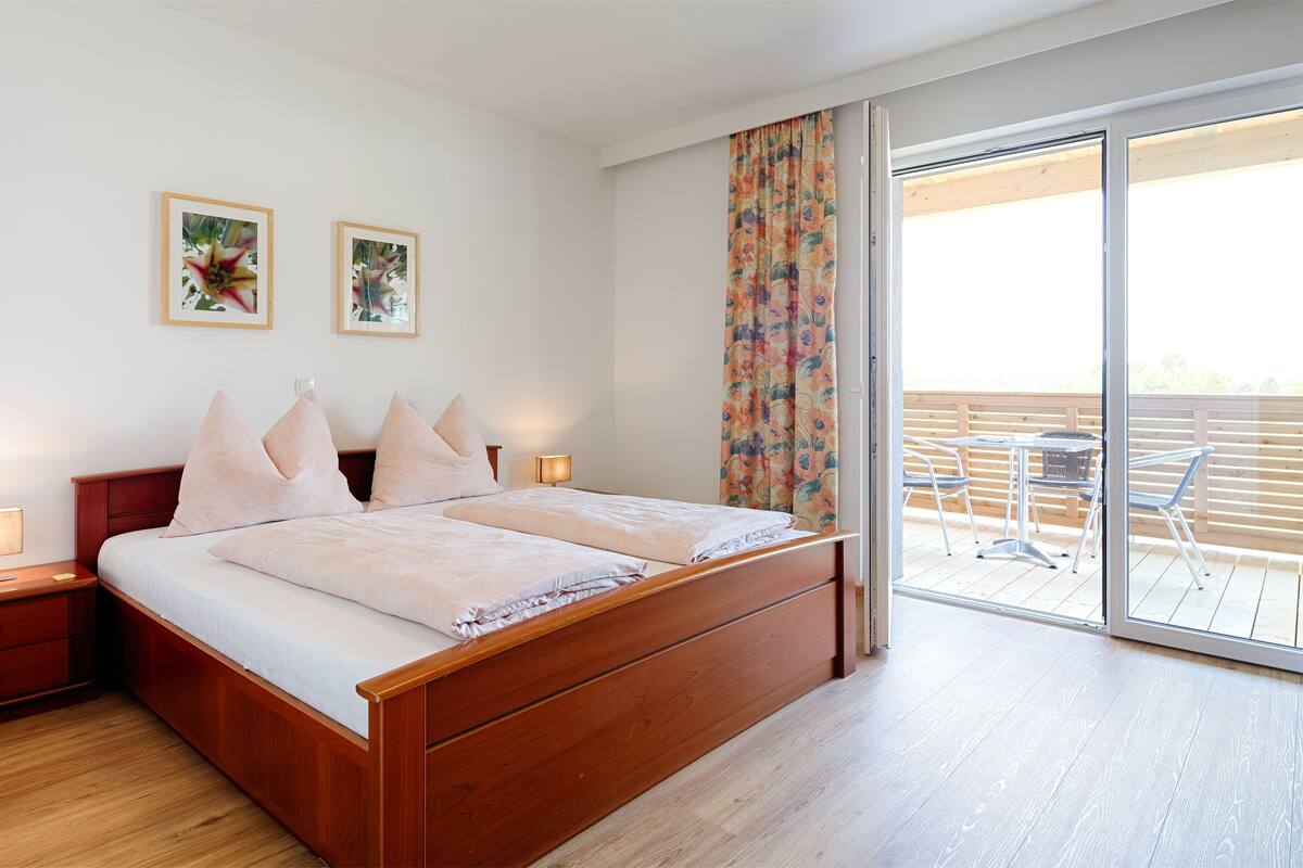 Un dormitorio con cama de madera, dos mesitas de noche, una mesita auxiliar, una ventana con cortina y un balcón con mesa y sillas.