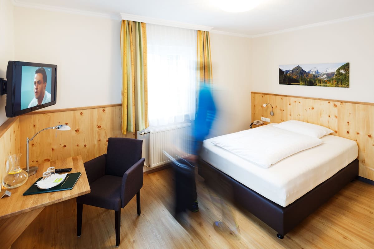 Habitación de hotel con cama, escritorio con lámpara, silla, televisor y ventana con cortinas.