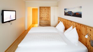 A hotel room with a wooden headboard, a flat-screen TV mounted on the wall, a bed with white linens, and a painting of mountains on the wall. - Pension Jageredt (Nussbach)