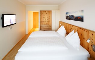 A hotel room with a wooden headboard, a flat-screen TV mounted on the wall, a bed with white linens, and a painting of mountains on the wall.