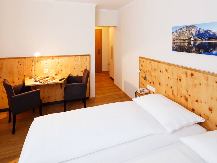 Habitación de hotel con cabecera de madera, cama con sábanas blancas, escritorio con dos sillas, teléfono y un cuadro con paisaje de montaña.