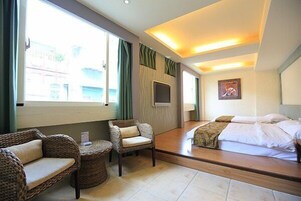 Habitación de hotel con una cama, dos sillas de mimbre, una mesita, televisor y un ventanal con cortinas.
