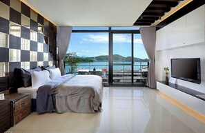 A modern bedroom with a large bed, a TV, and a balcony with a view of the sea.