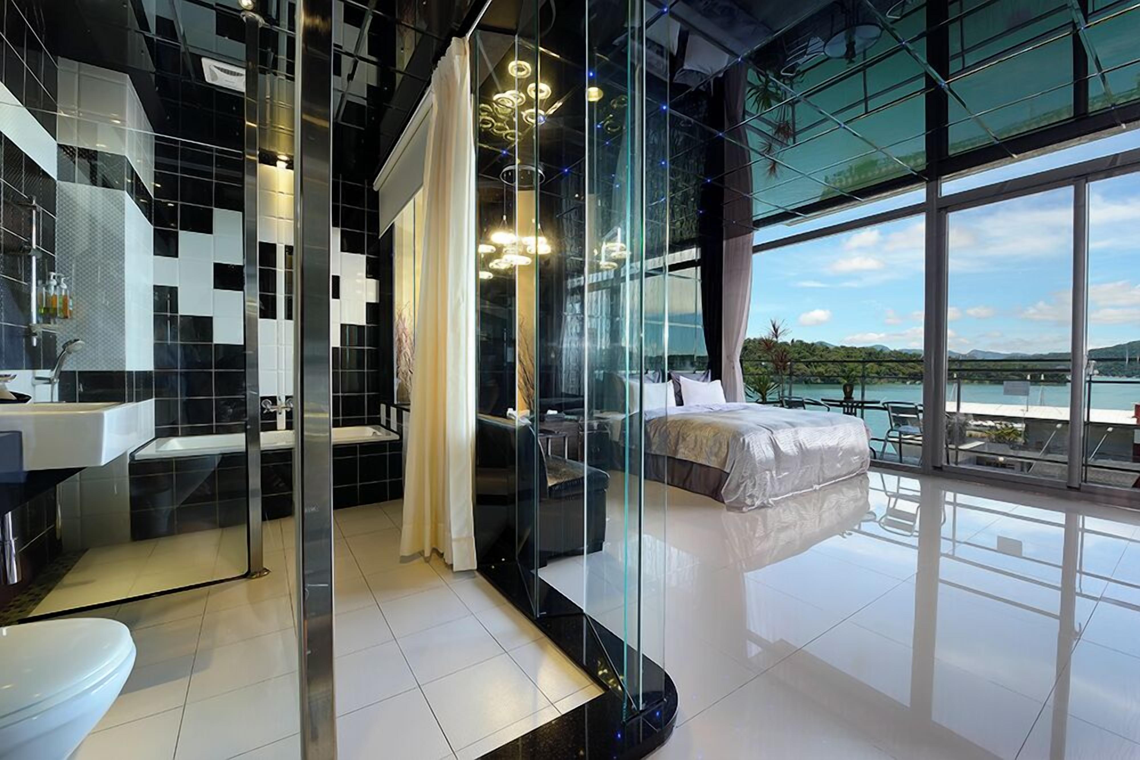 A modern hotel room with a glass partition, a large bed, a bathroom with a sink, and a view of the outdoors.