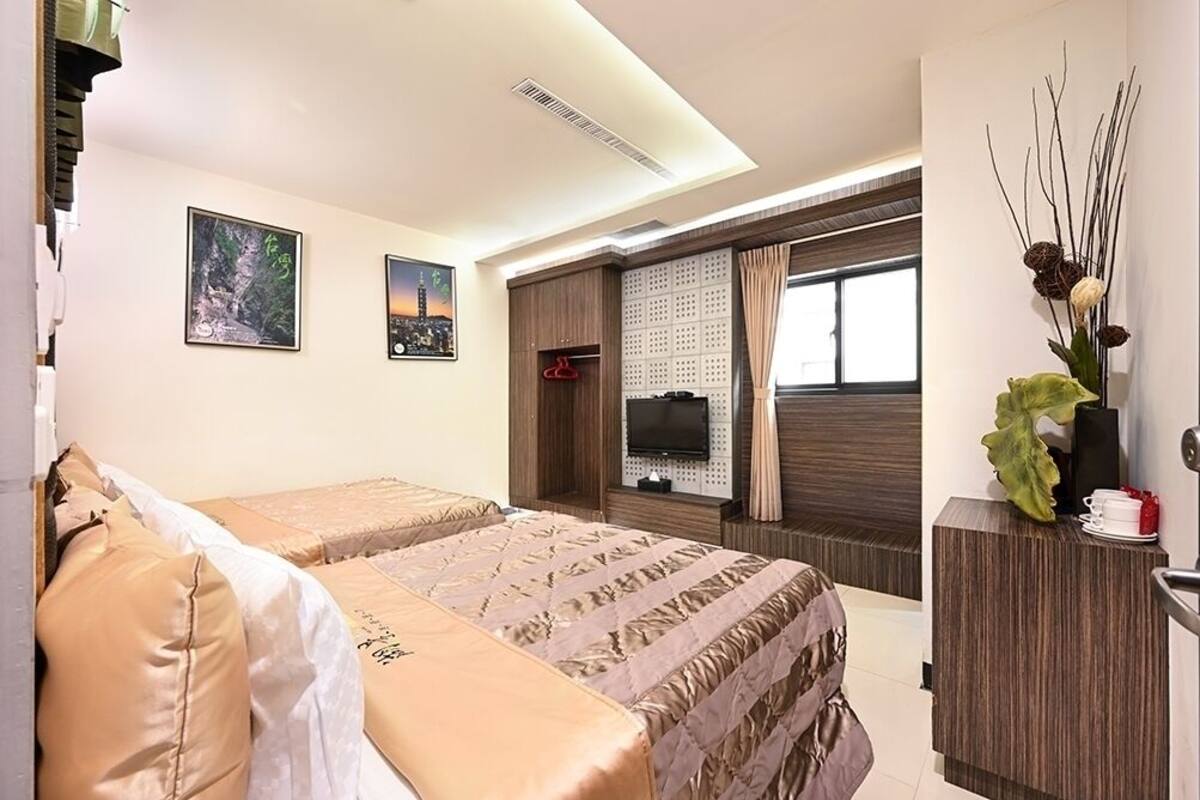 Ein Hotelzimmer mit zwei Betten, einem Fernseher, einem Schreibtisch mit einer Vase und Kunstwerken an den Wänden.