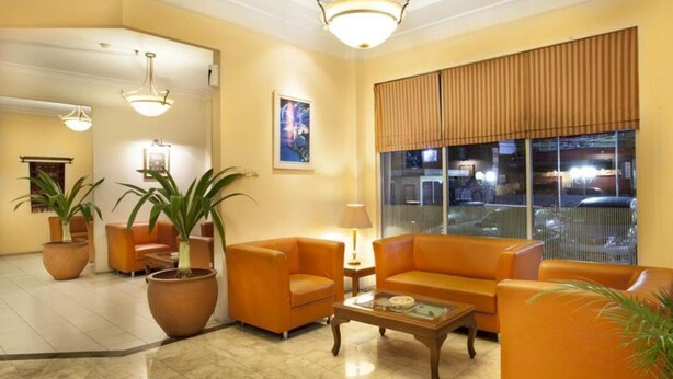 A lobby with orange sofas, a coffee table, potted plants, a large window with blinds, and a chandelier. - Nalendra (Bandung)