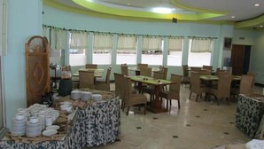 A dining area with tables and chairs, a buffet table with stacked plates, and a window with curtains. - Nalendra (Bandung)