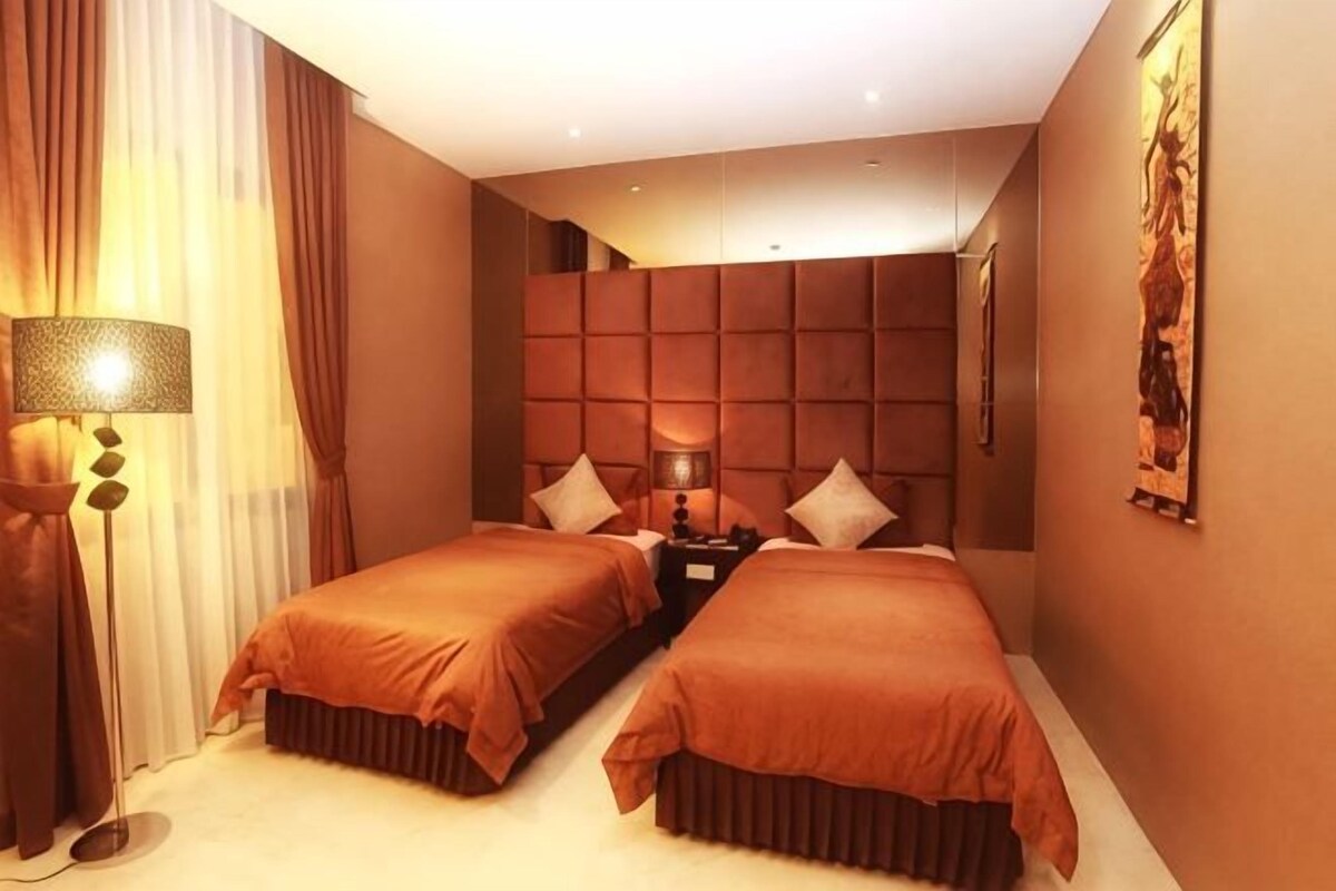 A hotel room with two beds, a large padded headboard, a bedside table with a lamp, and a wall-mounted artwork.