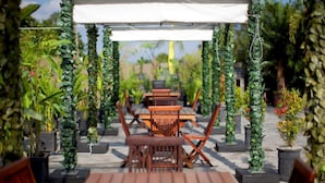 Un patio al aire libre con mesas y sillas de madera, rodeado de follaje y plantas en macetas.