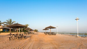 A beach with wooden picnic tables and thatched umbrellas, a clear sky, and a calm sea in the background. - Barracuda Beach Resort (Al Rafaah)