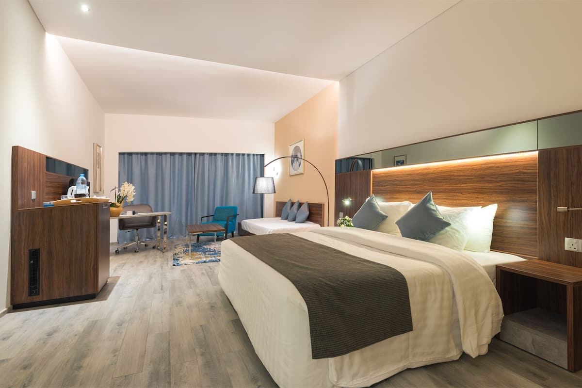 A hotel room with a large bed, a desk with a chair, a small table with a vase, and a window with curtains.