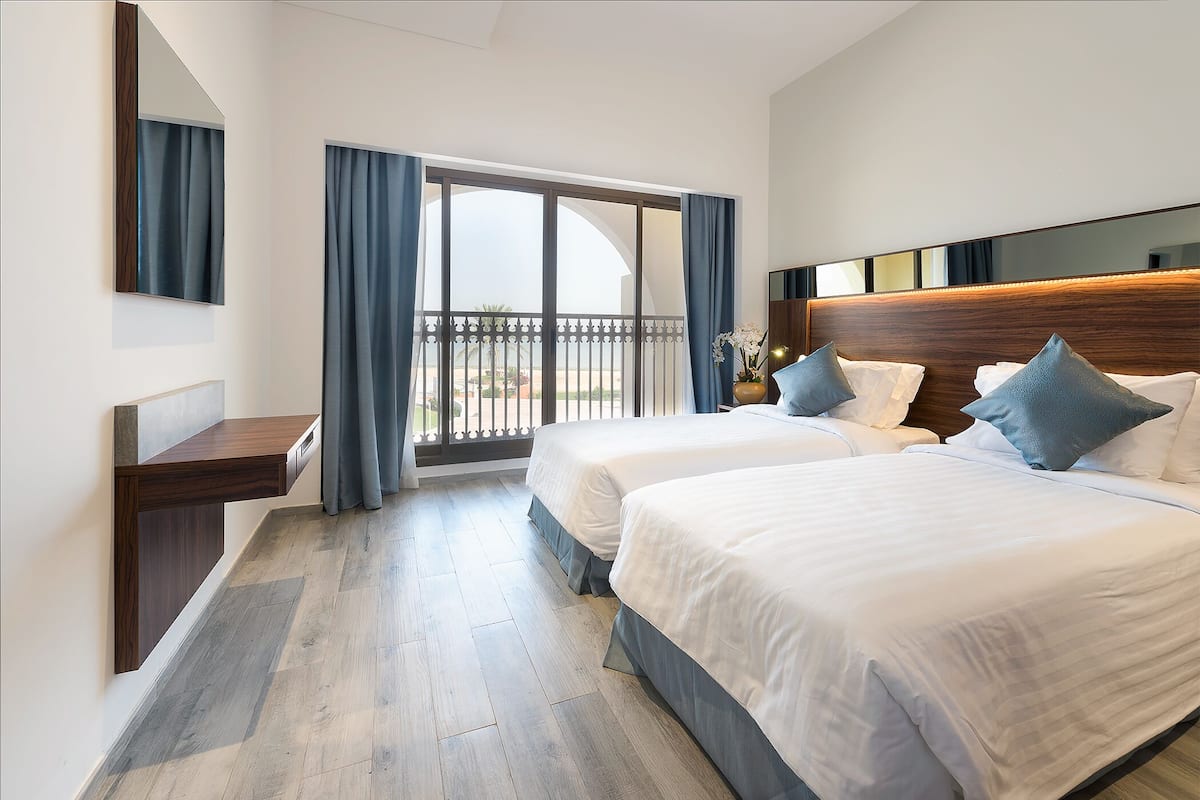 A hotel room with two beds, a wooden headboard, a large window with curtains, and a balcony.