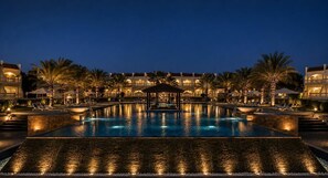 Uma piscina grande e bem iluminada, com um coreto no centro, rodeada por palmeiras e edifícios de hotel.