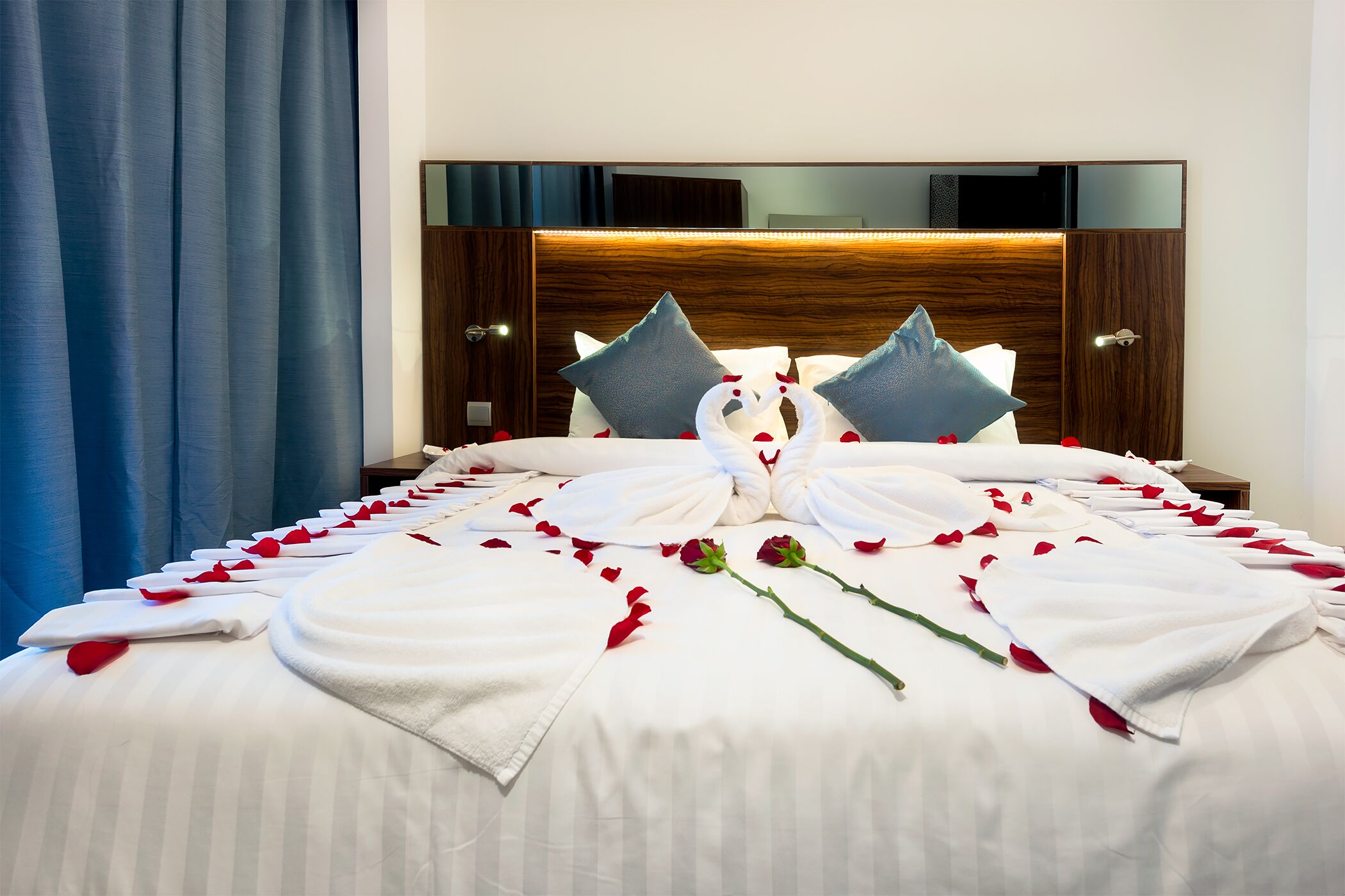 Un lit bien fait, décoré de pétales de roses et agrémenté d’un bouquet de roses, avec une tête de lit en bois et un grand miroir au-dessus du lit.