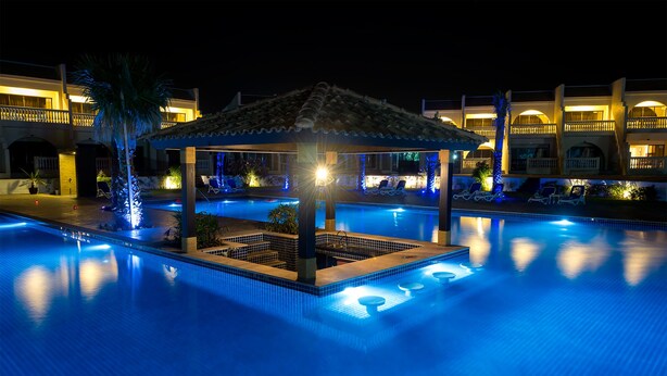 A nighttime swimming pool with a gazebo, lounge chairs, and a well-lit building in the background. - Barracuda Beach Resort (Al Rafaah)