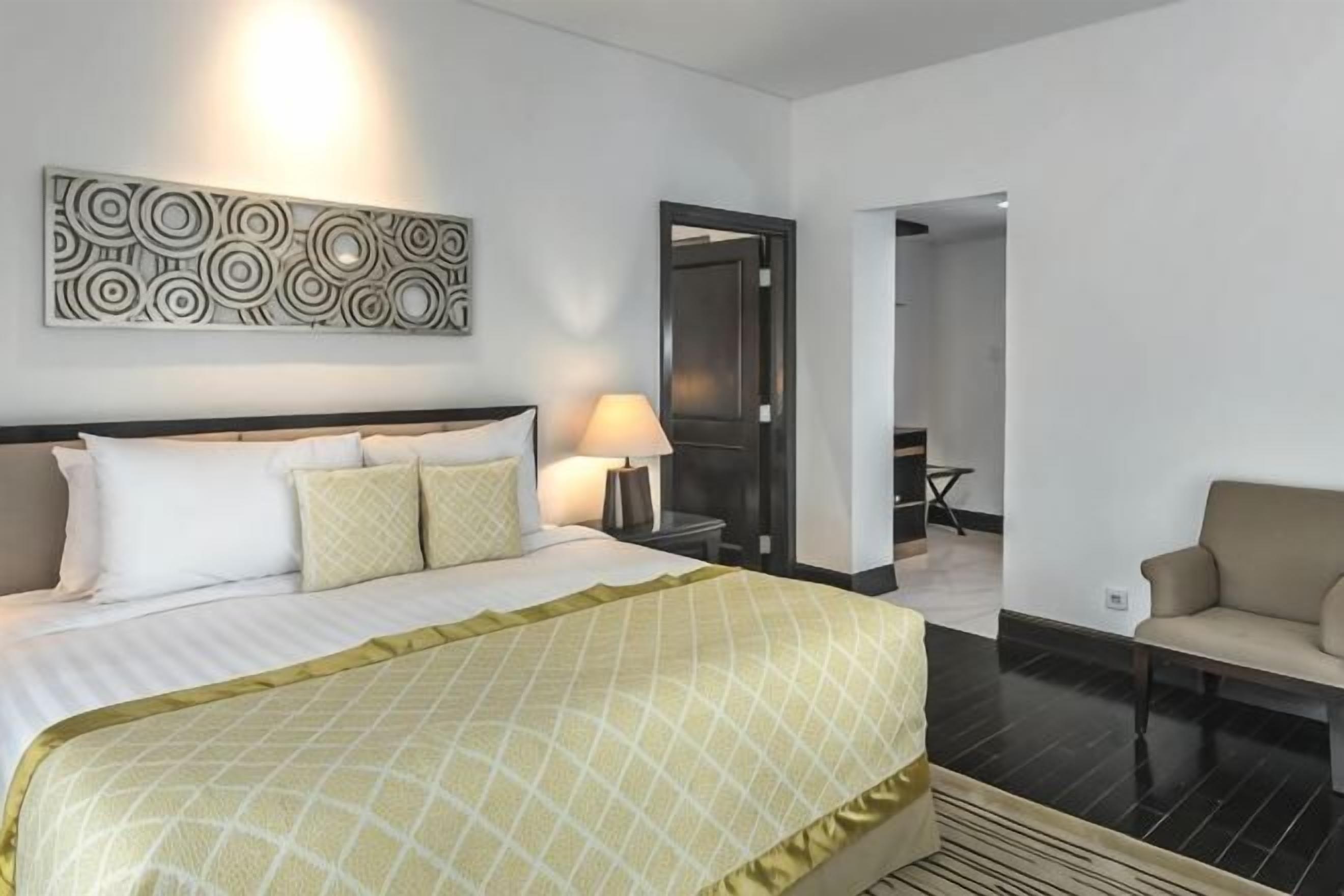 A hotel room with a bed, a chair, a lamp, and a door leading to another room.