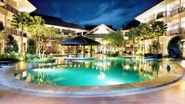 Vista nocturna de un patio de hotel con piscina, palmeras y un pabellón.