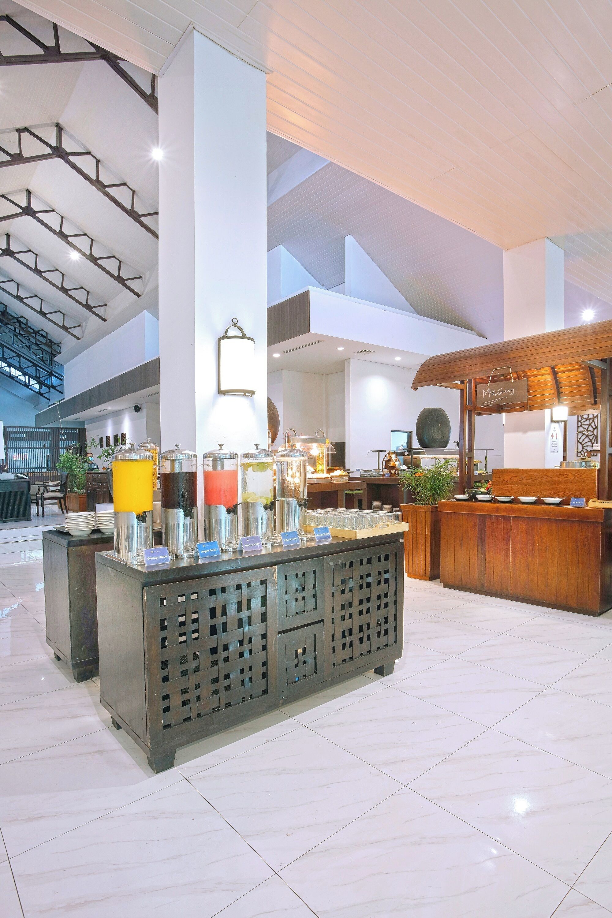 Un moderno vestíbulo de hotel con una barra de desayuno central, una recepción y un área de estar con mesas y sillas.