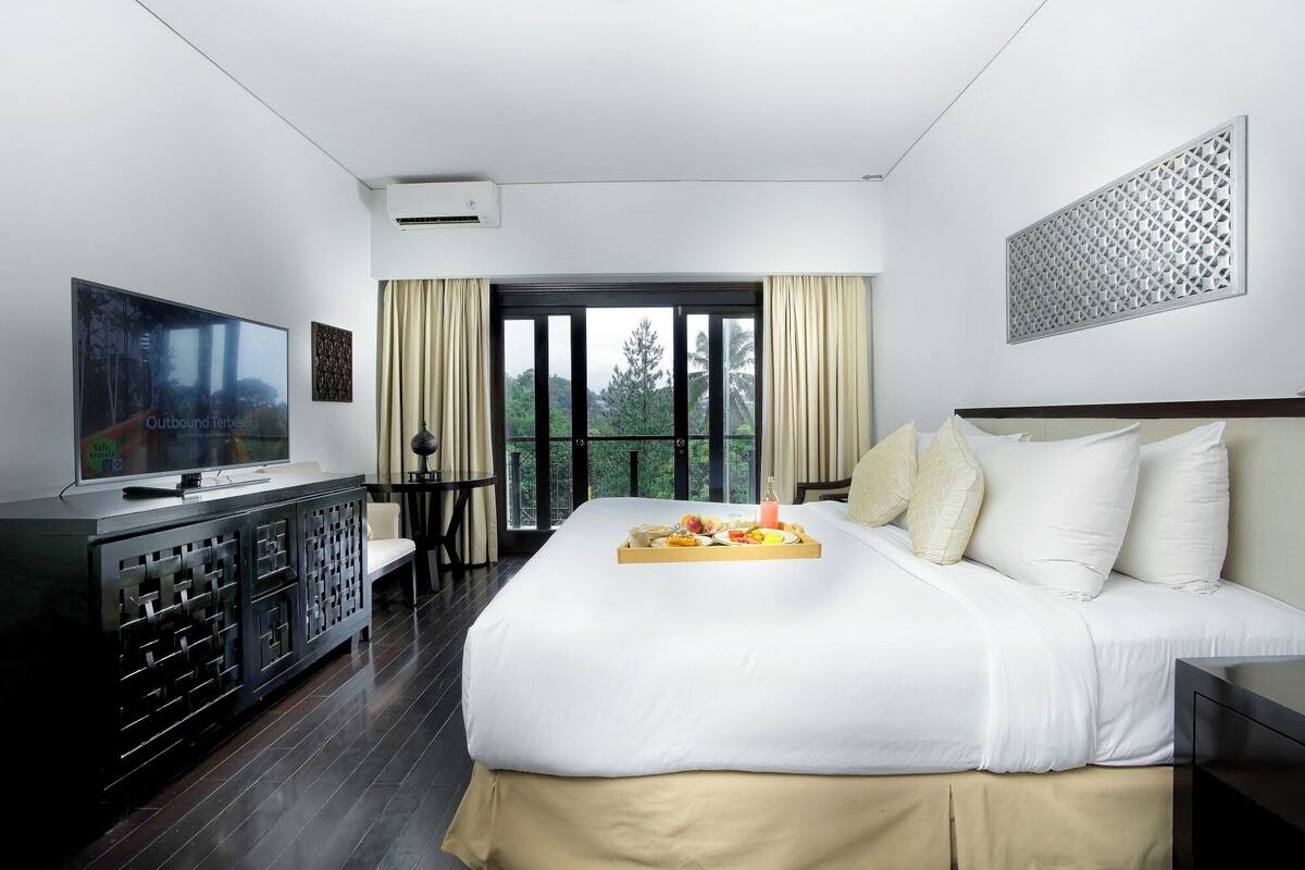 Une chambre d’hôtel avec un lit, une télévision, un balcon avec des rideaux et un plateau de petit-déjeuner posé sur le lit.