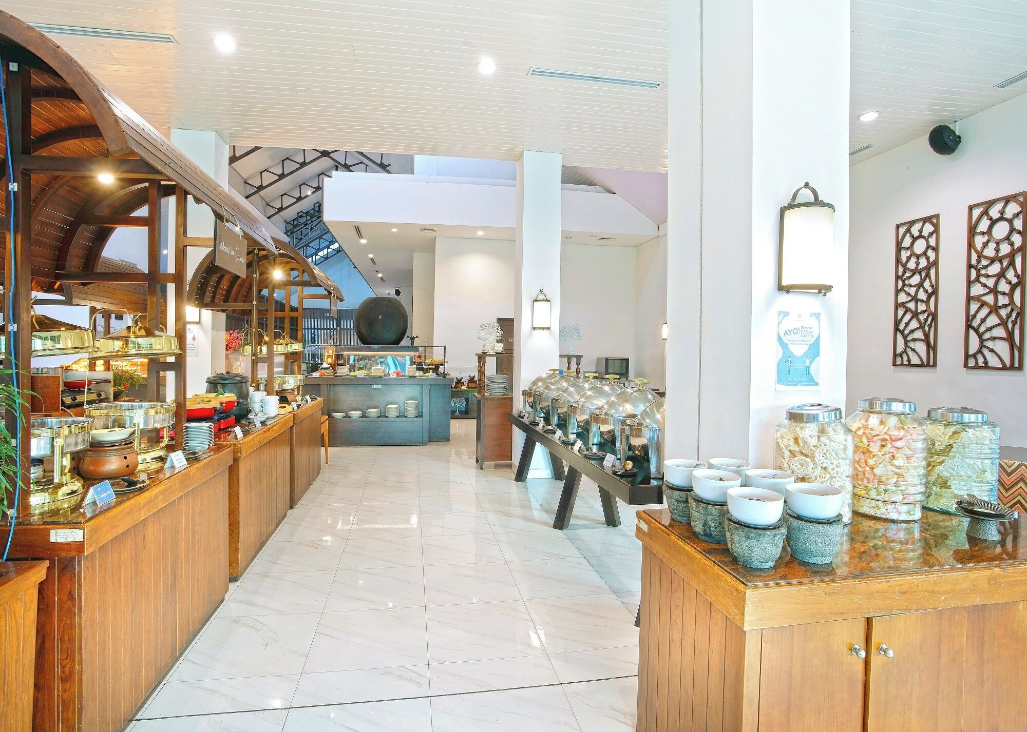 Um hall de hotel com um balcão de recepção central, uma área de buffet com recipientes de comida e uma escadaria que leva a um andar superior.