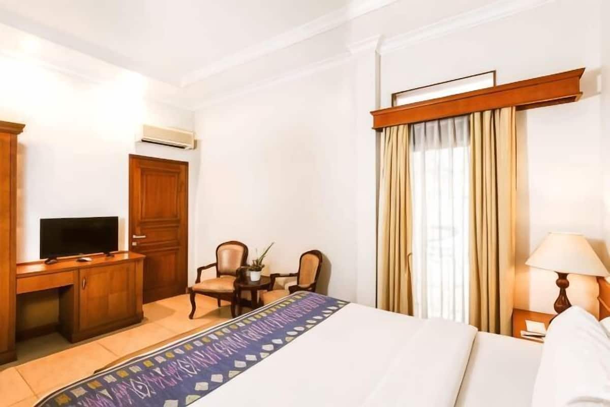 A hotel room with a bed, a desk with a television, two chairs, a lamp, and a window with curtains.
