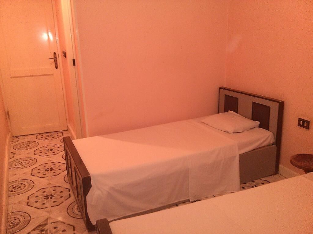 Una habitación pequeña con cama individual, cabecera de madera, colcha blanca y suelo estampado.