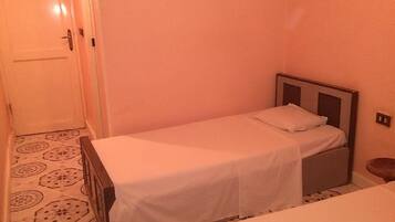 Una habitación pequeña con cama individual, cabecera de madera, colcha blanca y suelo estampado.