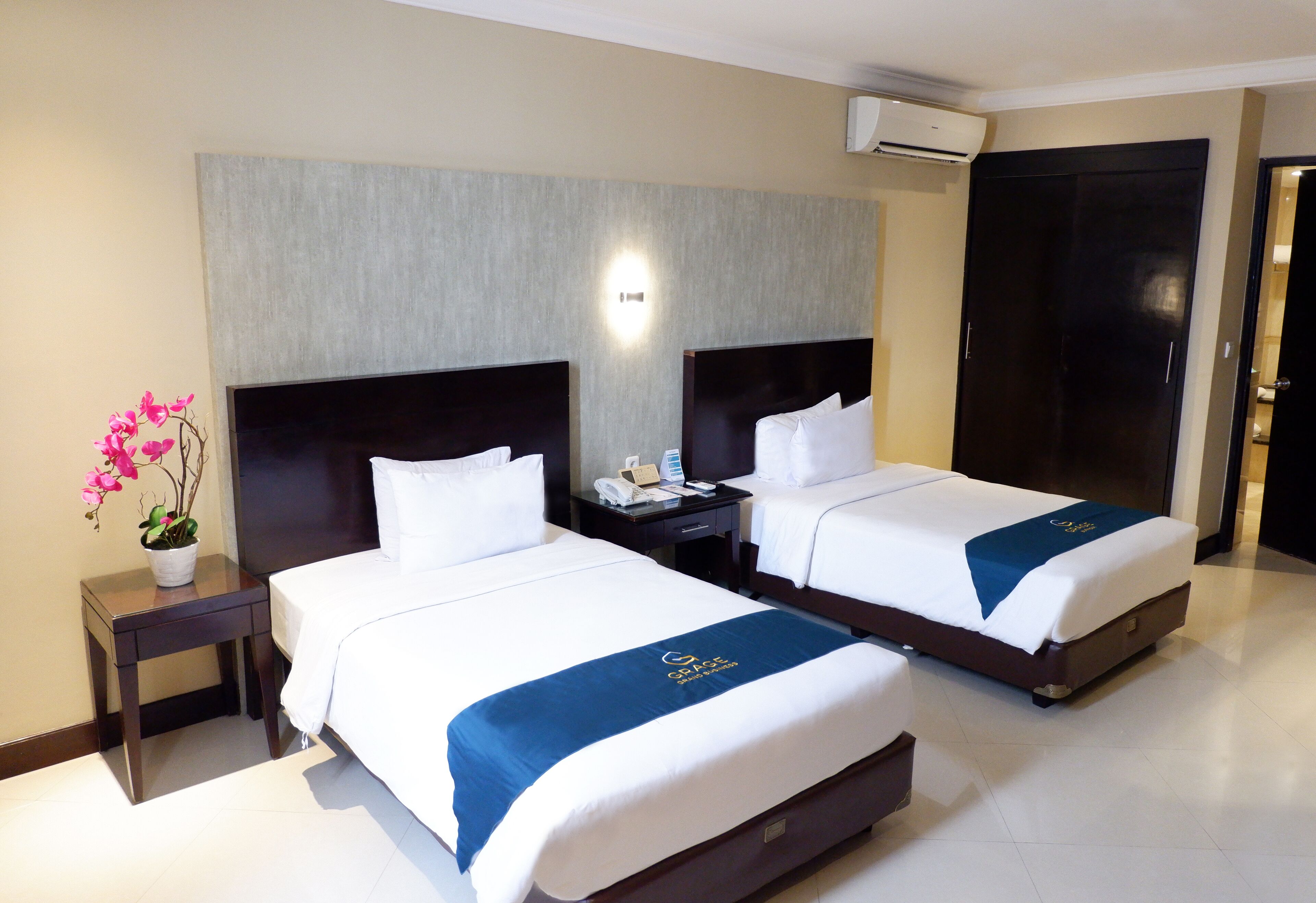 A hotel room with two beds, a nightstand, a potted plant, and a wall-mounted air conditioner.