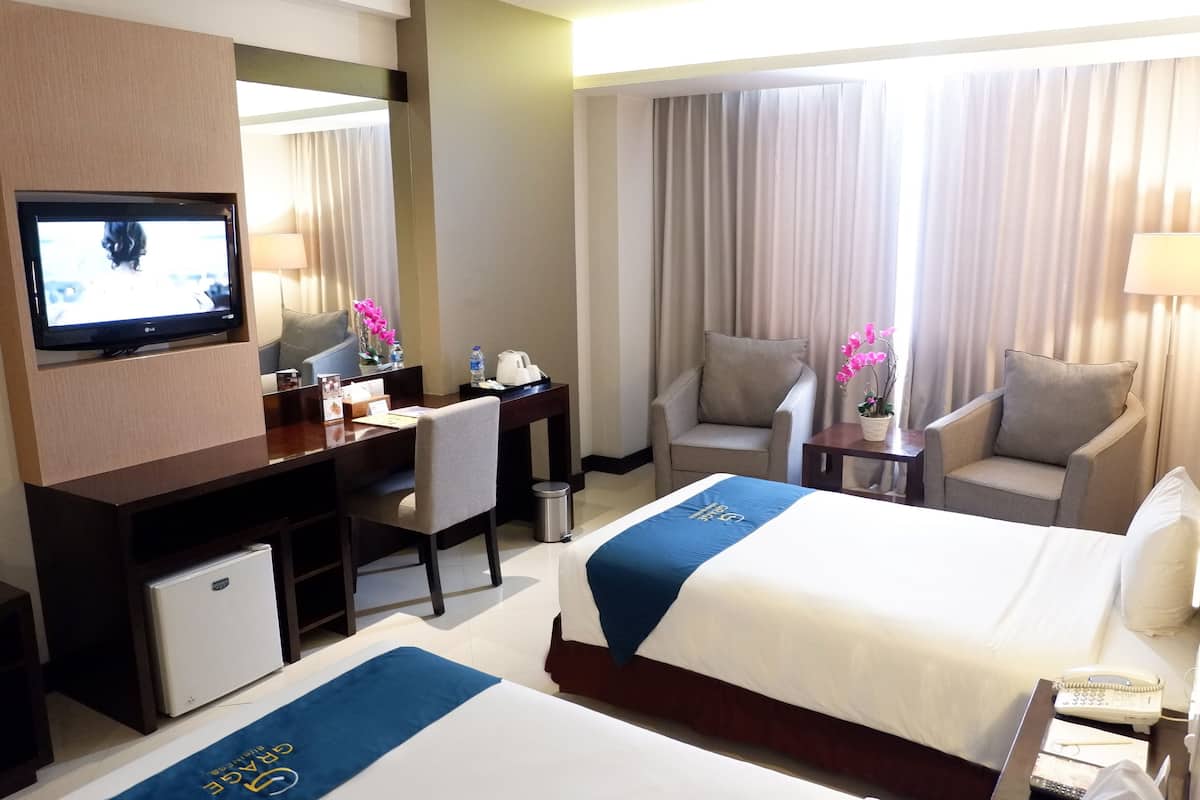 Habitación de hotel con dos camas, un escritorio con televisor, una silla, una mesita de noche con lámpara y una ventana con cortinas.