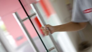 Une personne tenant un poteau métallique, vraisemblablement un dispositif de sécurité, dans un couloir avec des portes en verre et un mur rose.