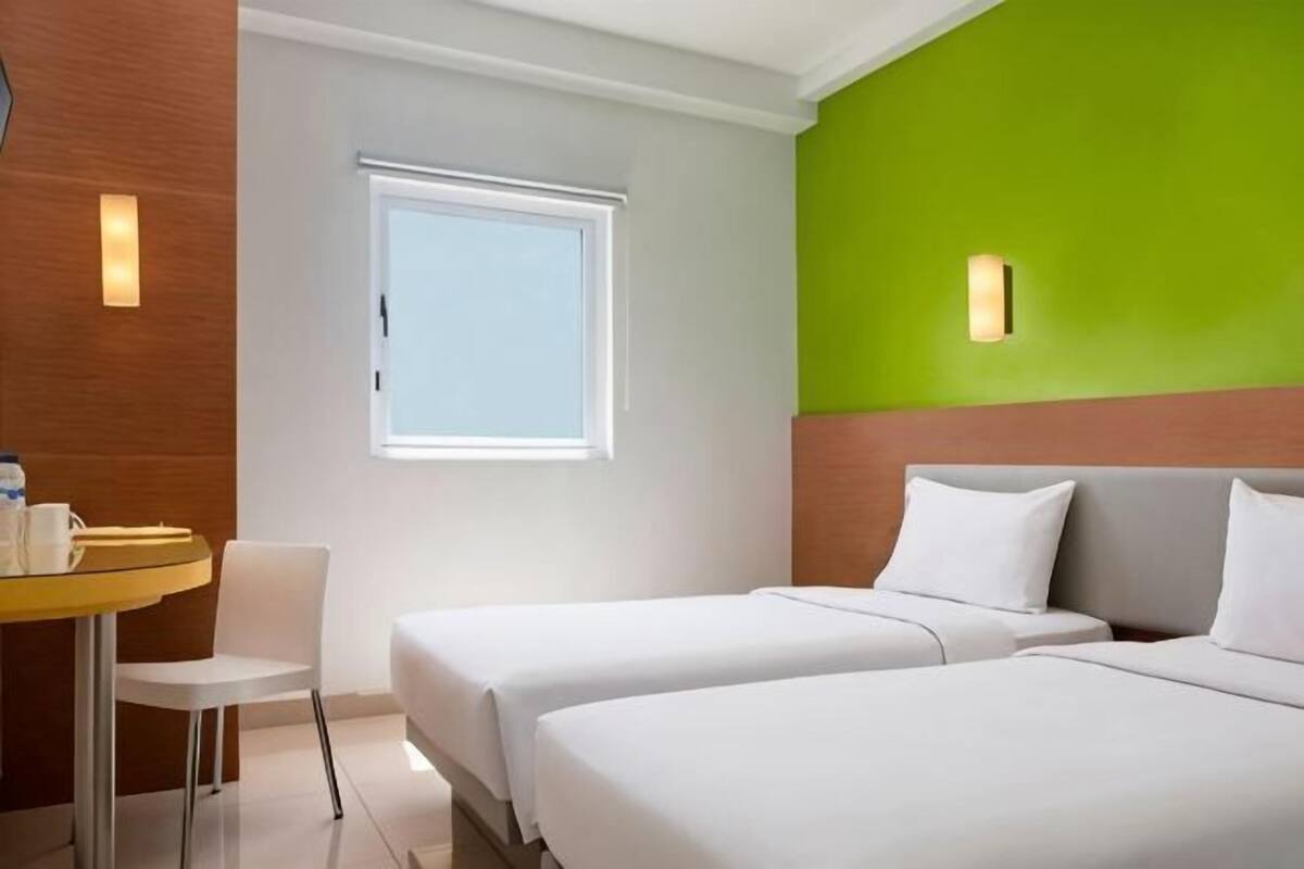 A hotel room with two beds, a small table with a chair, a window, and a green accent wall.