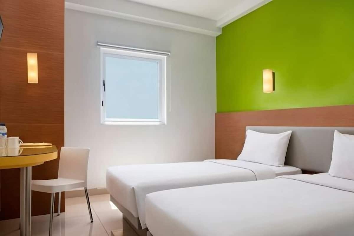 A hotel room with two beds, a small table with a chair, a window, and a green accent wall.