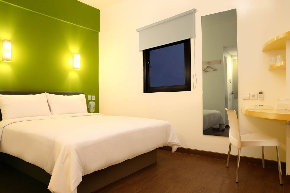 A hotel room with a bed, a desk, a chair, a mirror, and a window with a view of the outside.