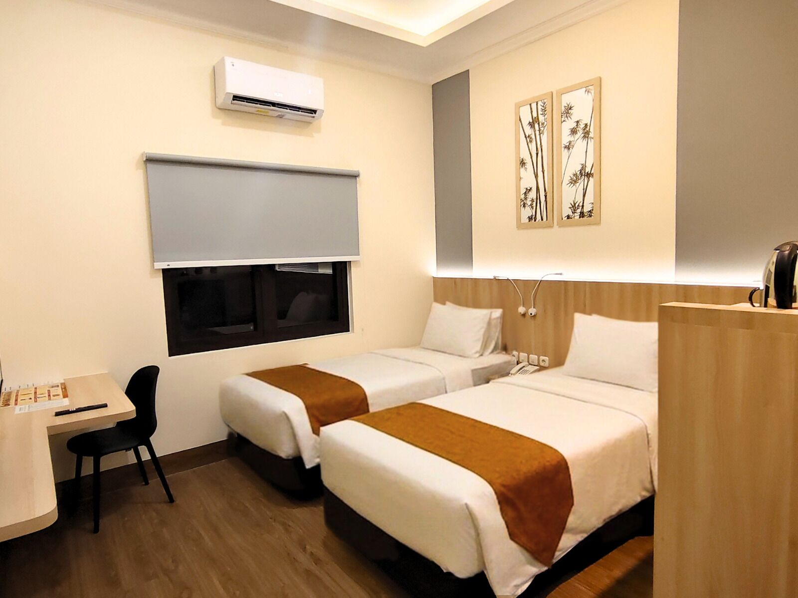 A hotel room with two beds, a desk, a chair, an air conditioning unit, and artwork on the wall.