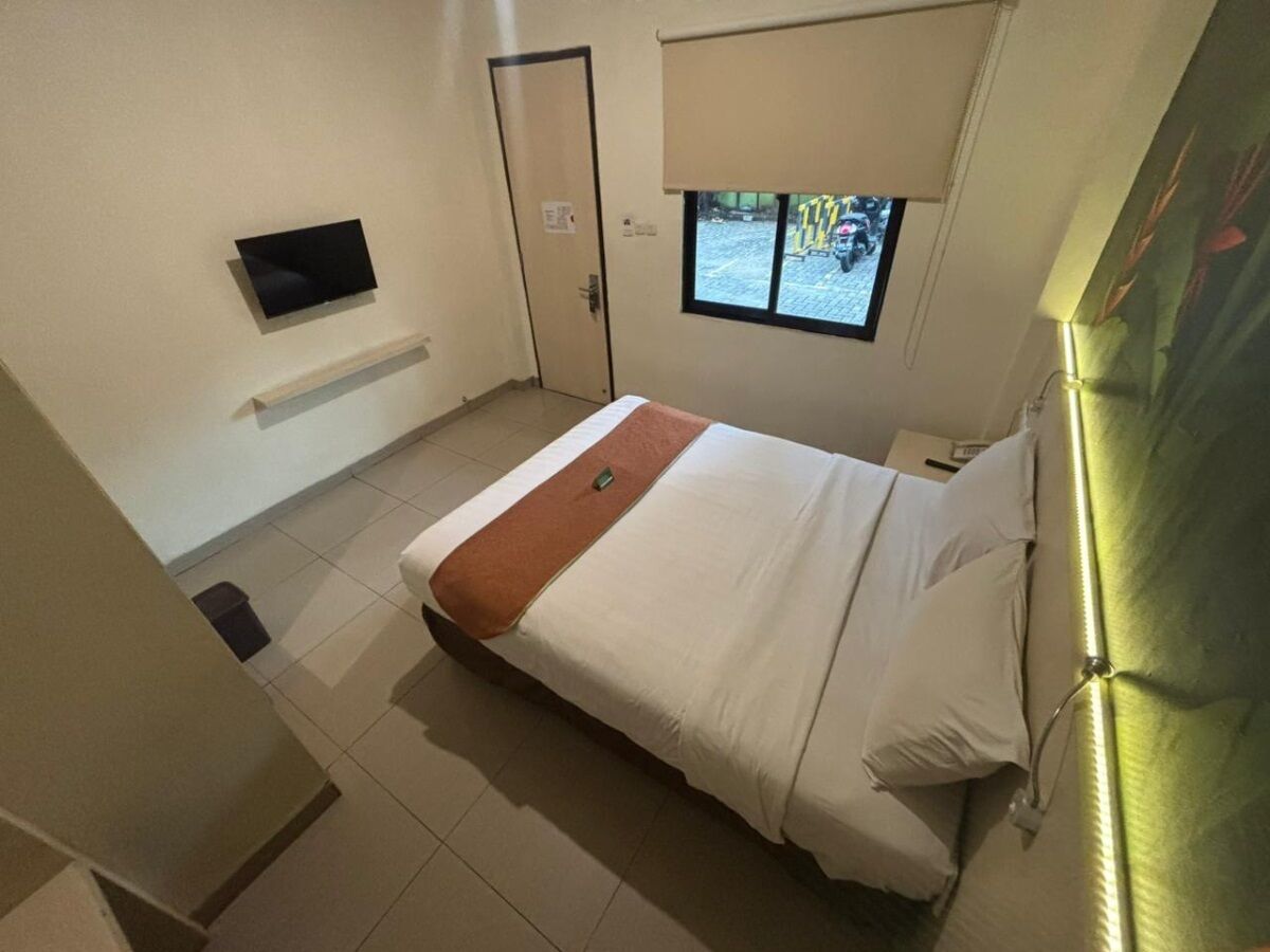 Una habitación de hotel con cama, televisor montado en la pared, ventana con vista a la calle y una puerta.