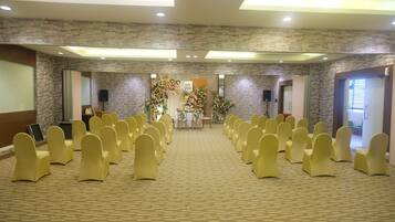 Un salón de conferencias con hileras de sillas amarillas, un escenario con adornos florales y un fondo de pared de piedra.