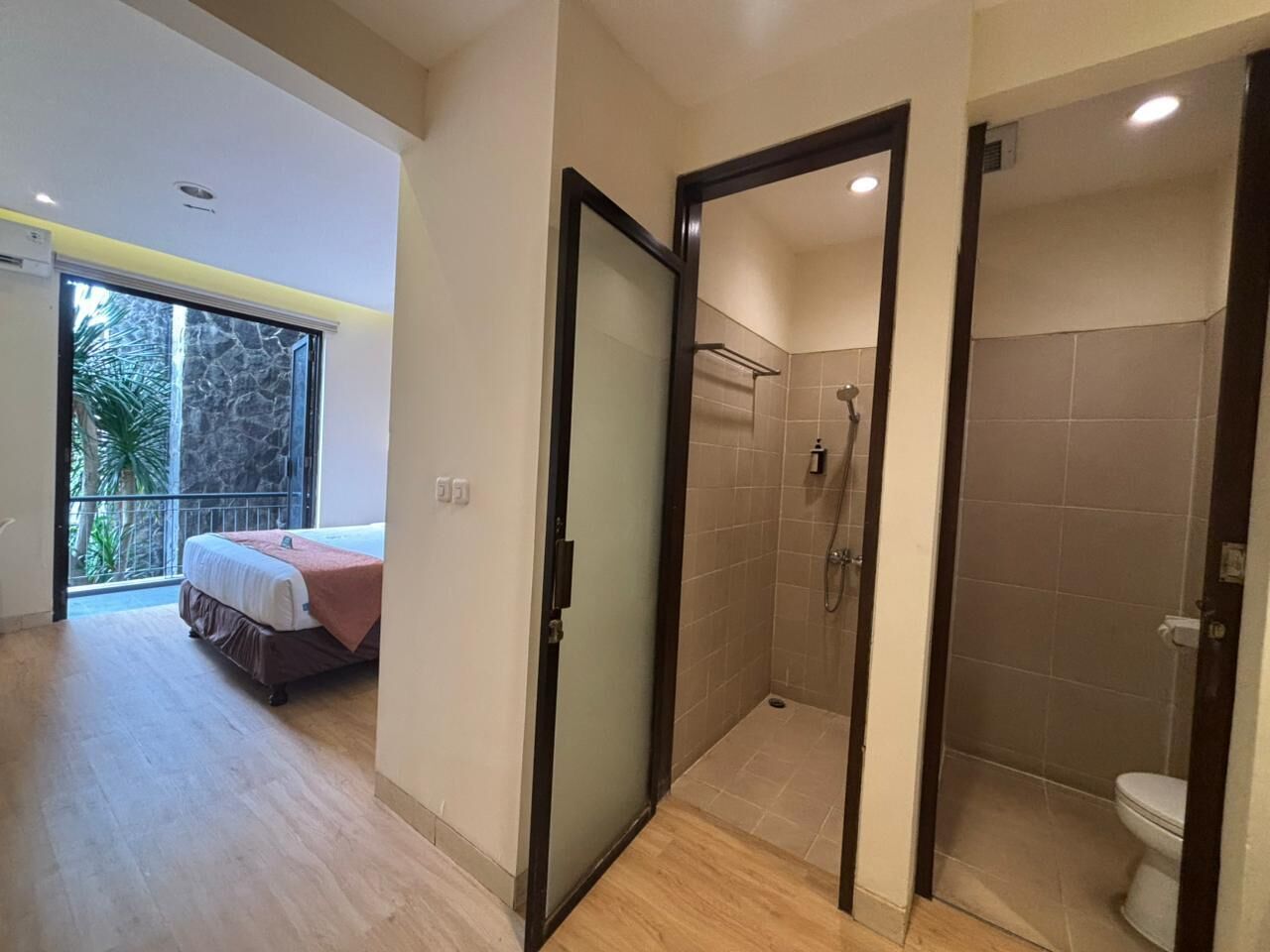 Ein modernes Hotelzimmer mit einem großen Bett, einer Duschkabine und einem Balkon mit Blick auf Palmen.