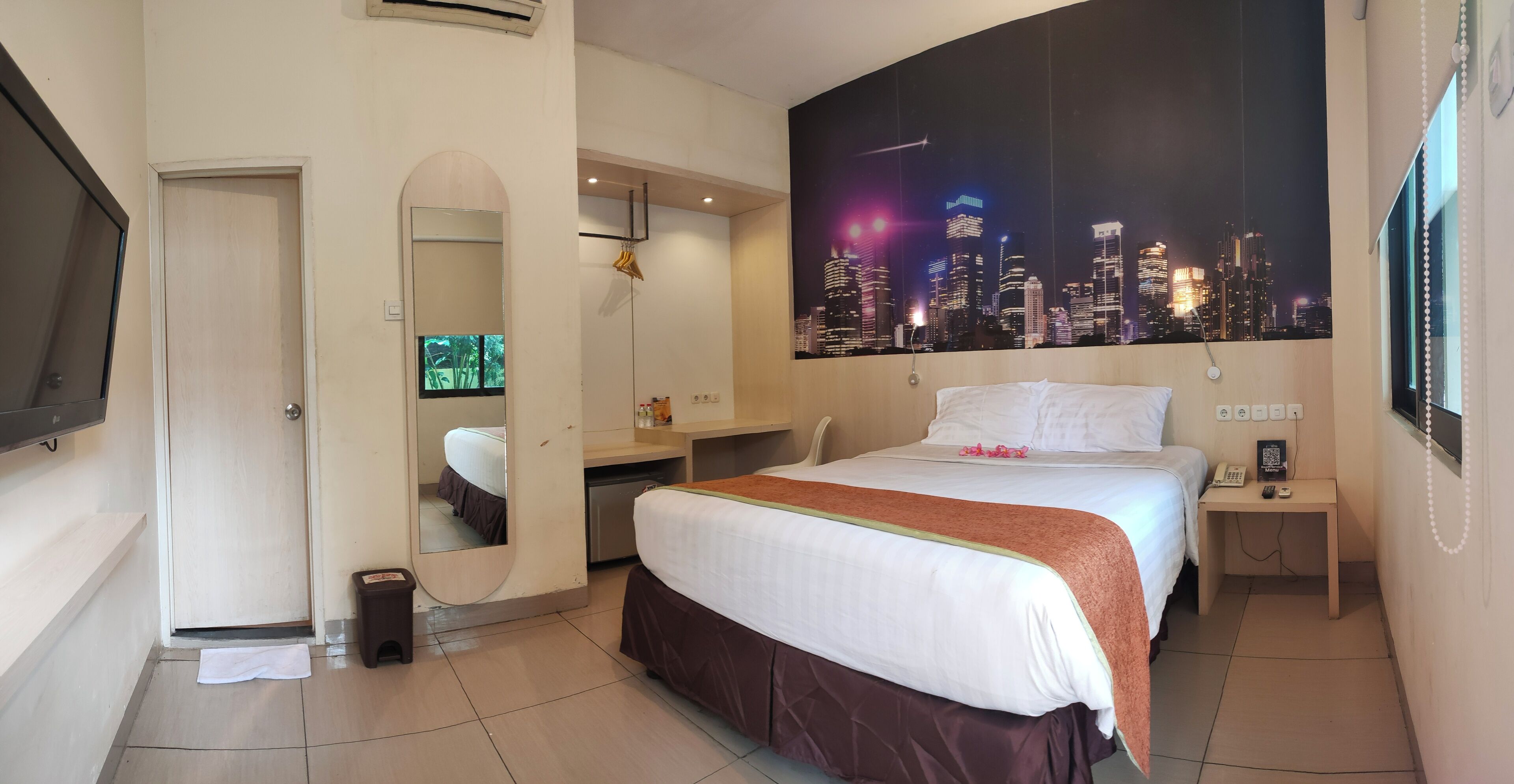A hotel room with a bed, a desk, a mirror, a nightstand, a telephone, and a cityscape view.