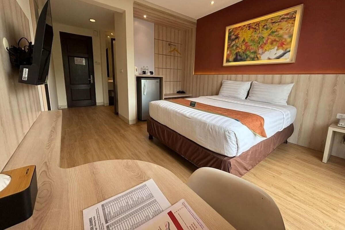 Habitación de hotel con cama, escritorio, silla, televisión, refrigerador y un cuadro en la pared.
