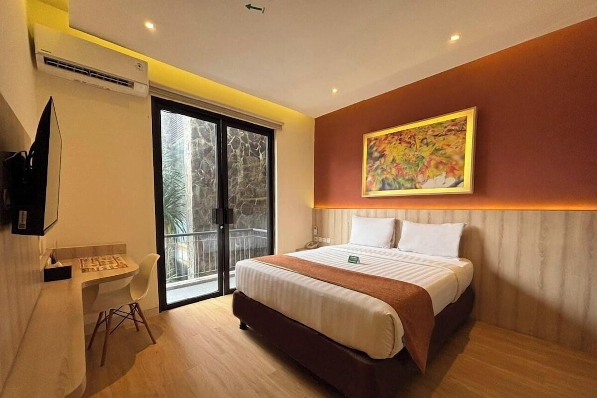 A modern hotel room with a bed, a desk with a telephone, a painting on the wall, and a sliding glass door leading to an outdoor area.