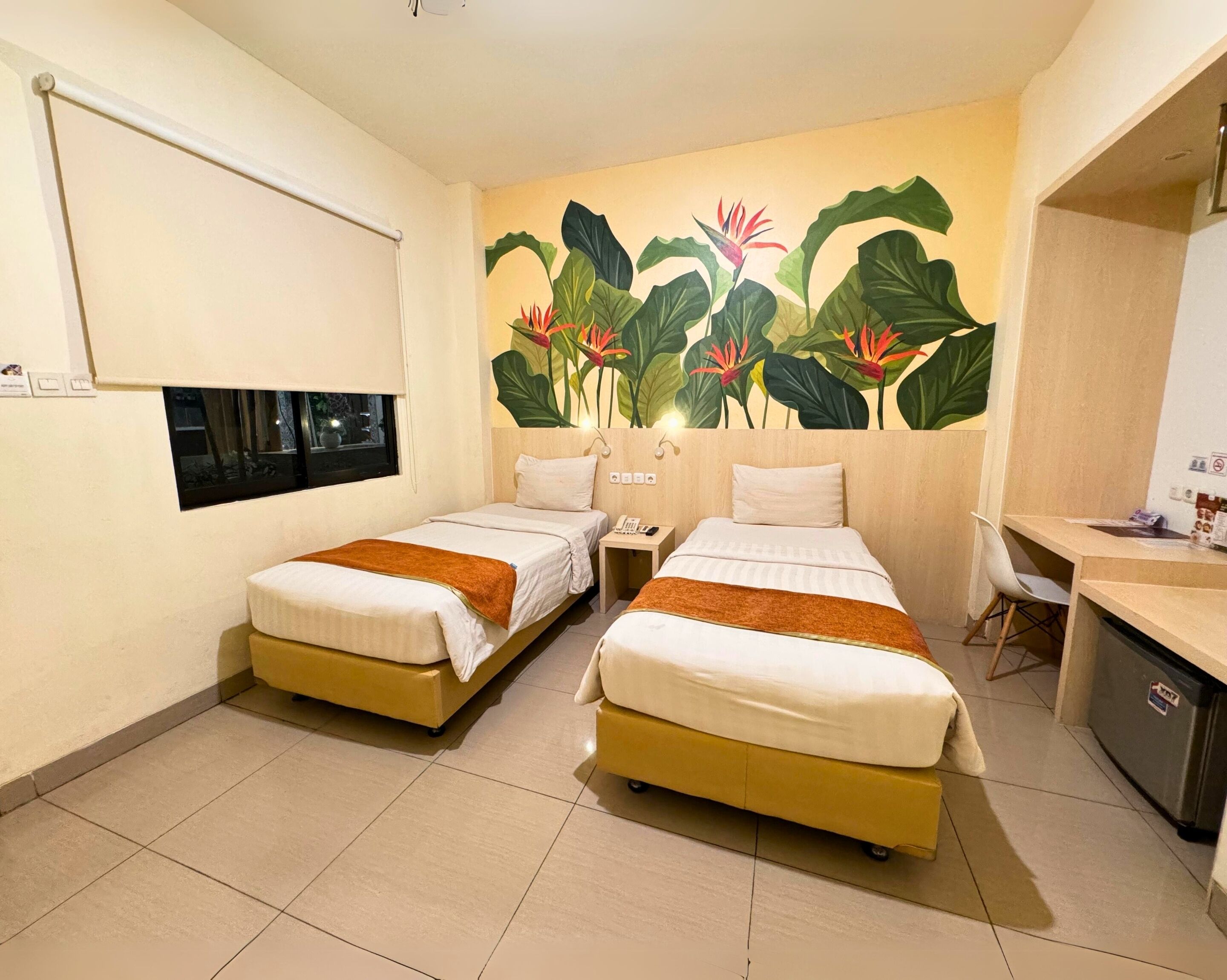 A hotel room with two beds, a desk, a chair, a small refrigerator, and a large wall mural of tropical plants.