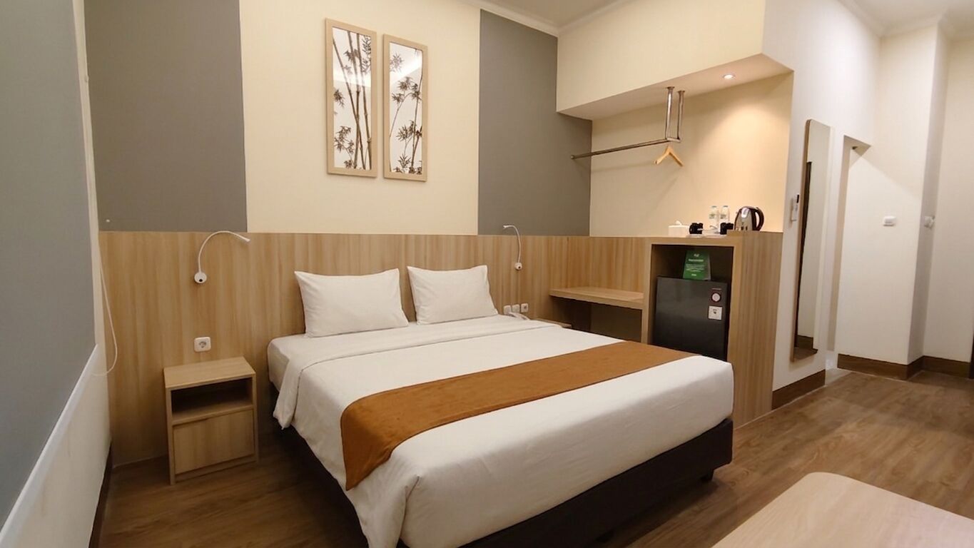 A hotel room with a bed, bedside tables, a small desk, a refrigerator, and a wall-mounted mirror.