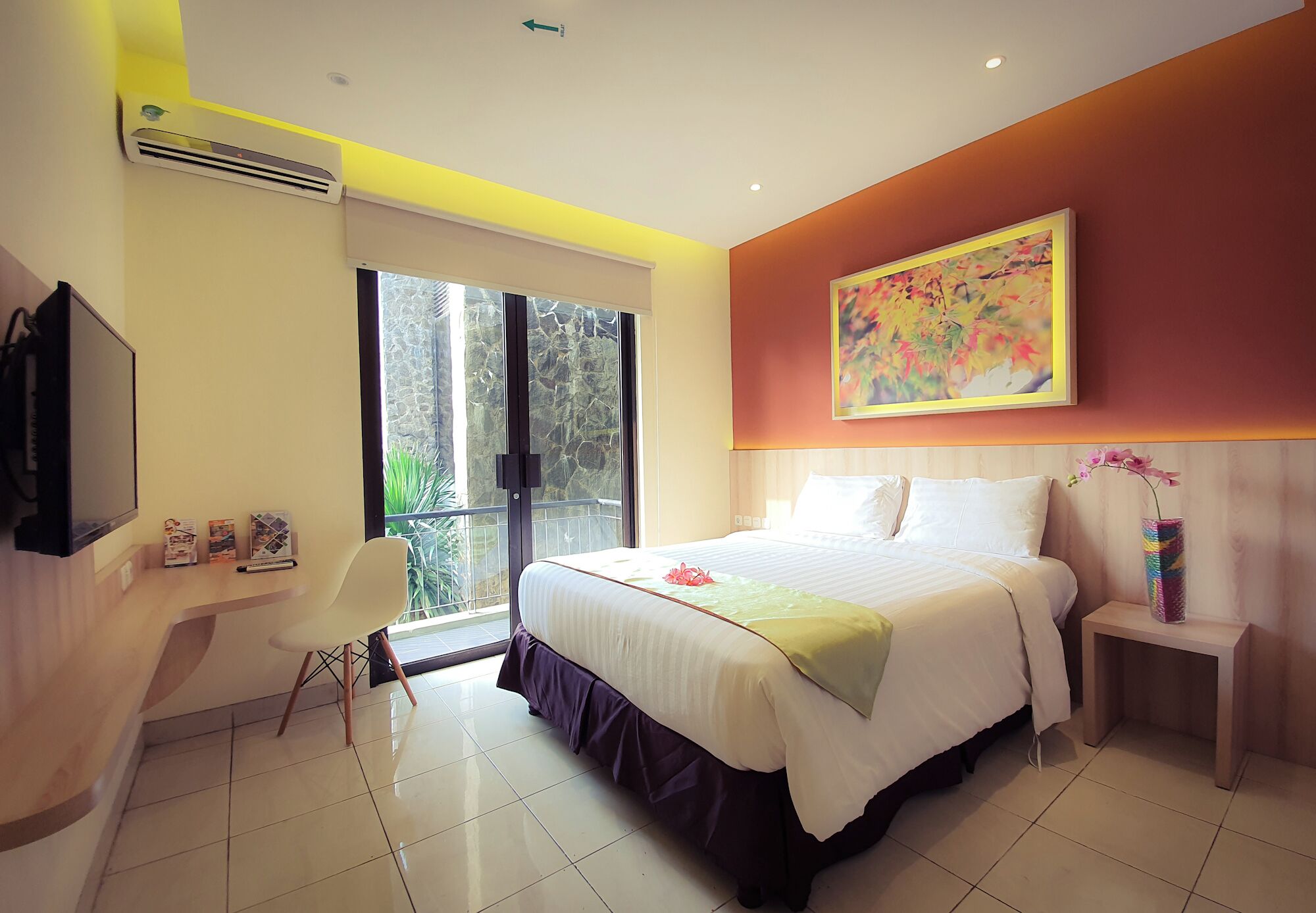 A modern hotel room with a large bed, a desk, a chair, a TV, and a window with a view of greenery.