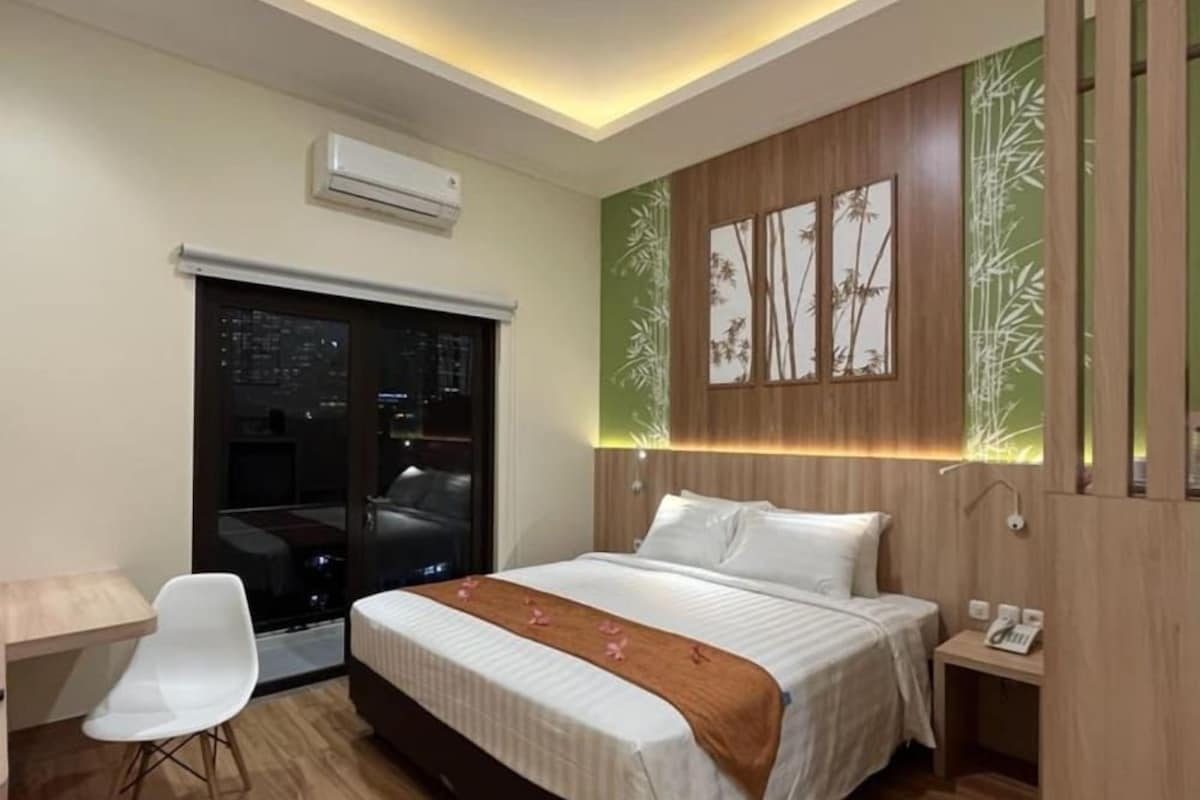 A modern hotel room with a bed, bedside table, chair, and a view of the city at night through a glass door.