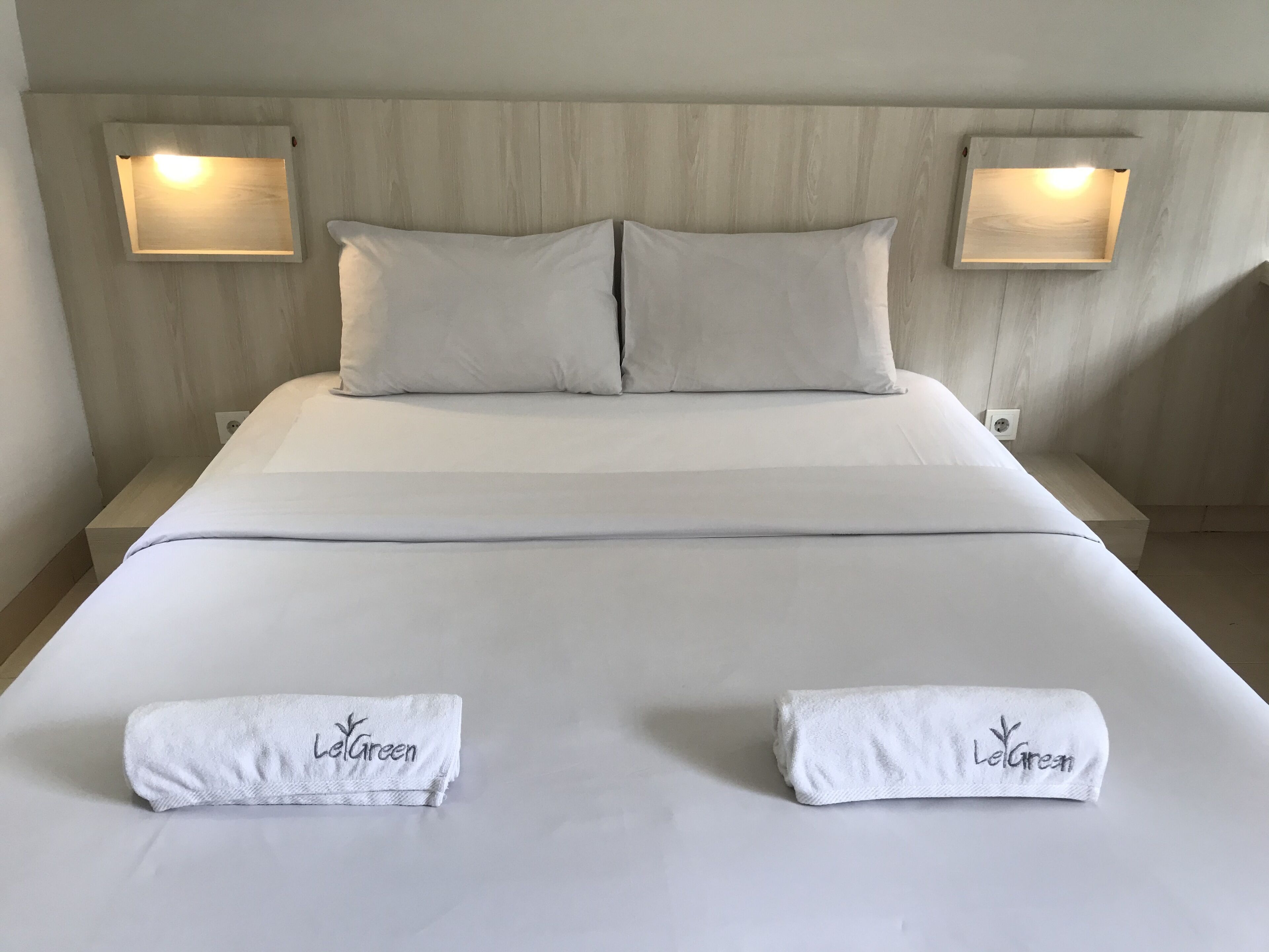 A neatly made bed with two towels labeled "LeGreen" and two wall-mounted lights on either side.