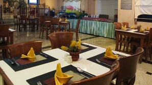 Uma área de refeições com mesas preparadas para quatro pessoas, cada uma com um guardanapo amarelo, saleiro e pimenteiro, e um pequeno vaso de planta.