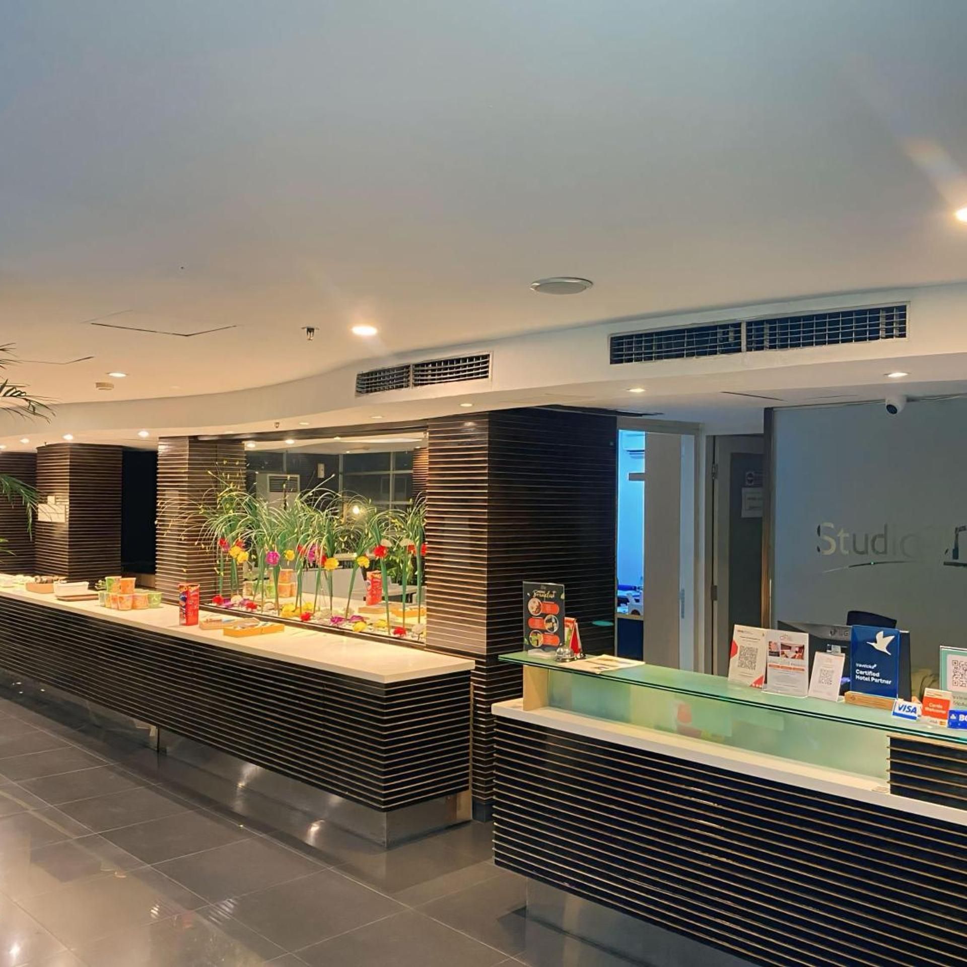 A hotel reception area with a modern design, featuring a reception desk, a plant display, and a sign indicating a partnership with a credit card company.